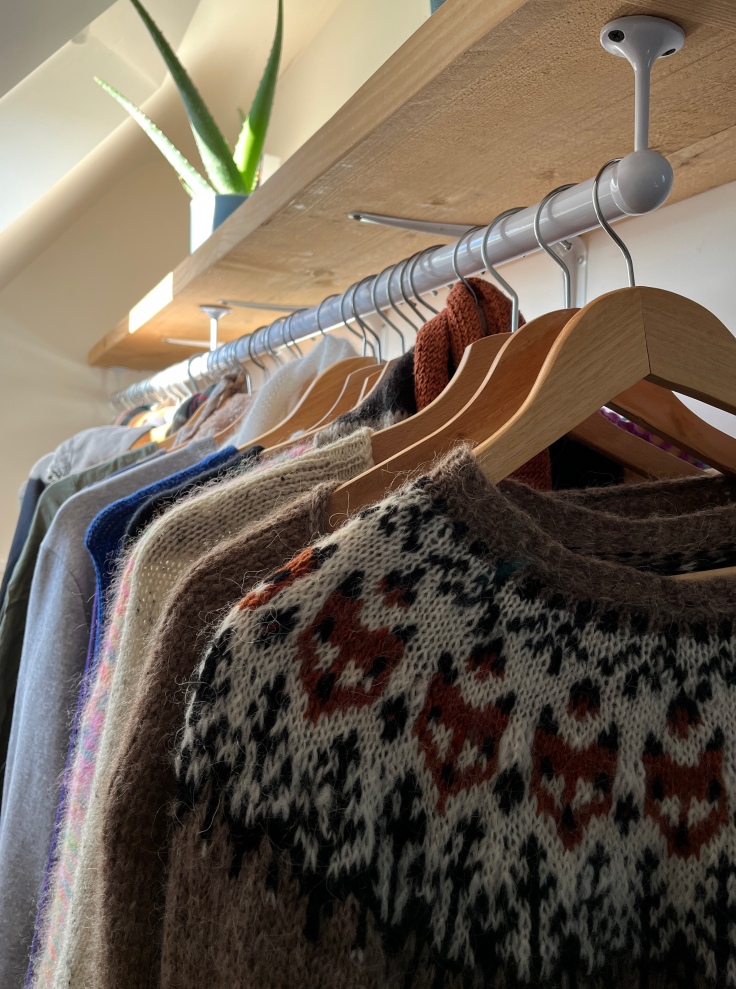 A selection of hand knitted sweaters hand on wooden hangers below a wooden shelf.