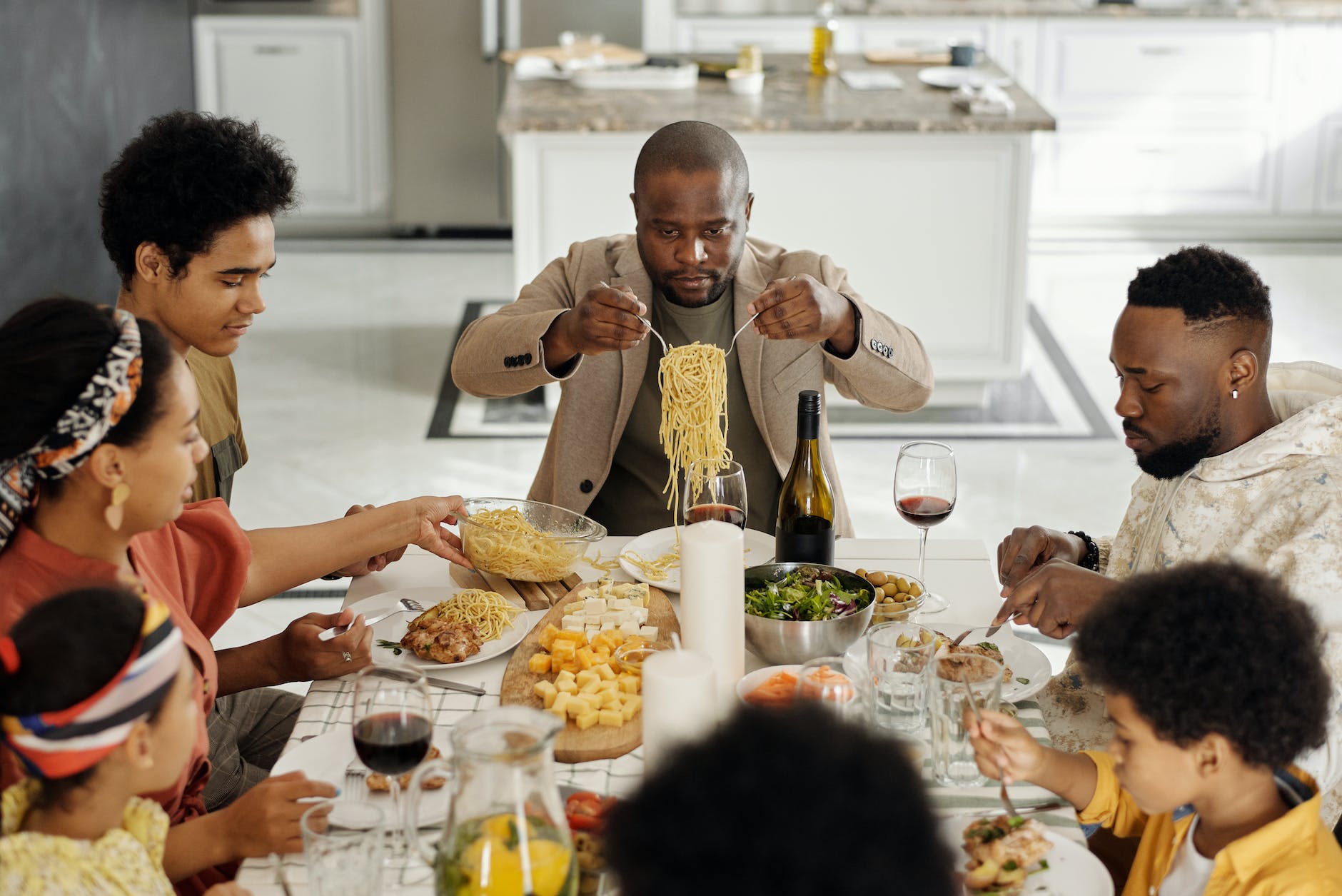 A family sat around a table eating a meal. A man sat at the head of the table holds up a serving on pasta. 