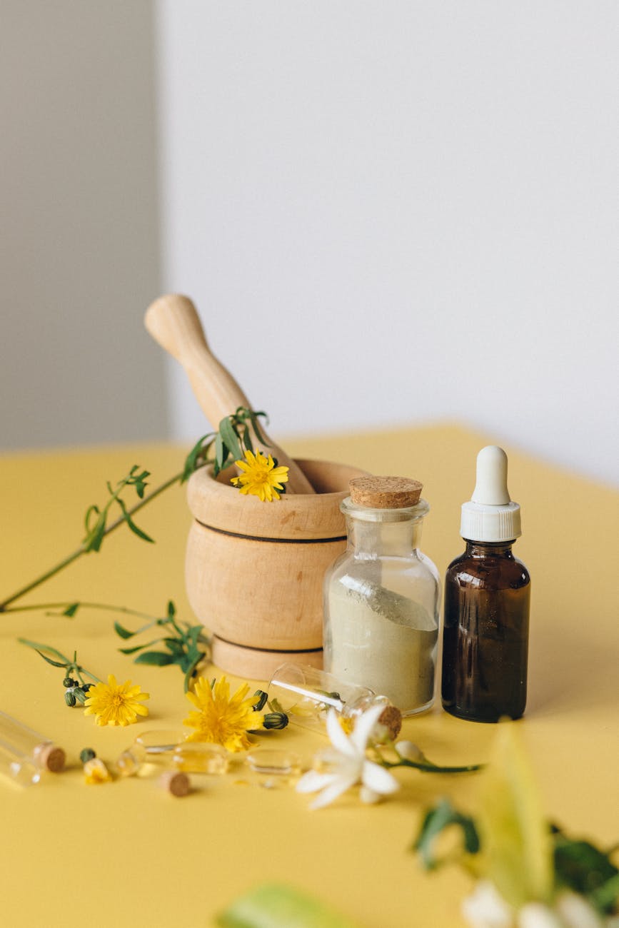 a selection of herbs and flowers and ointment container on a yellow table with a wooden pestle and mortar. 